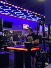 Person standing at a glowing console in a neon-lit gaming arcade with hexagonal ceiling lights