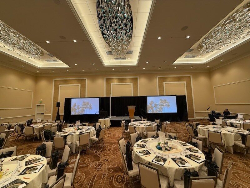 Banquet hall set for a conference, with round tables and two large projection screens at the front.