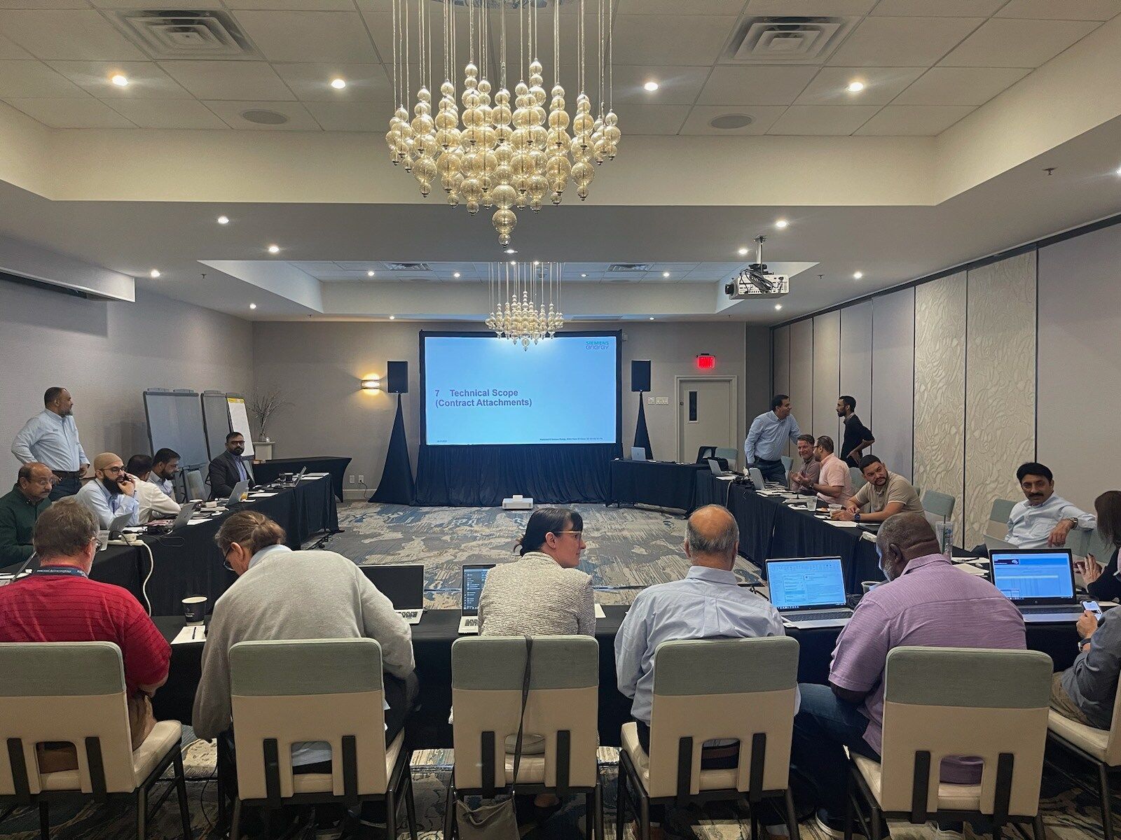 Conference room with people seated at tables facing a presentation screen and speaker at the front.