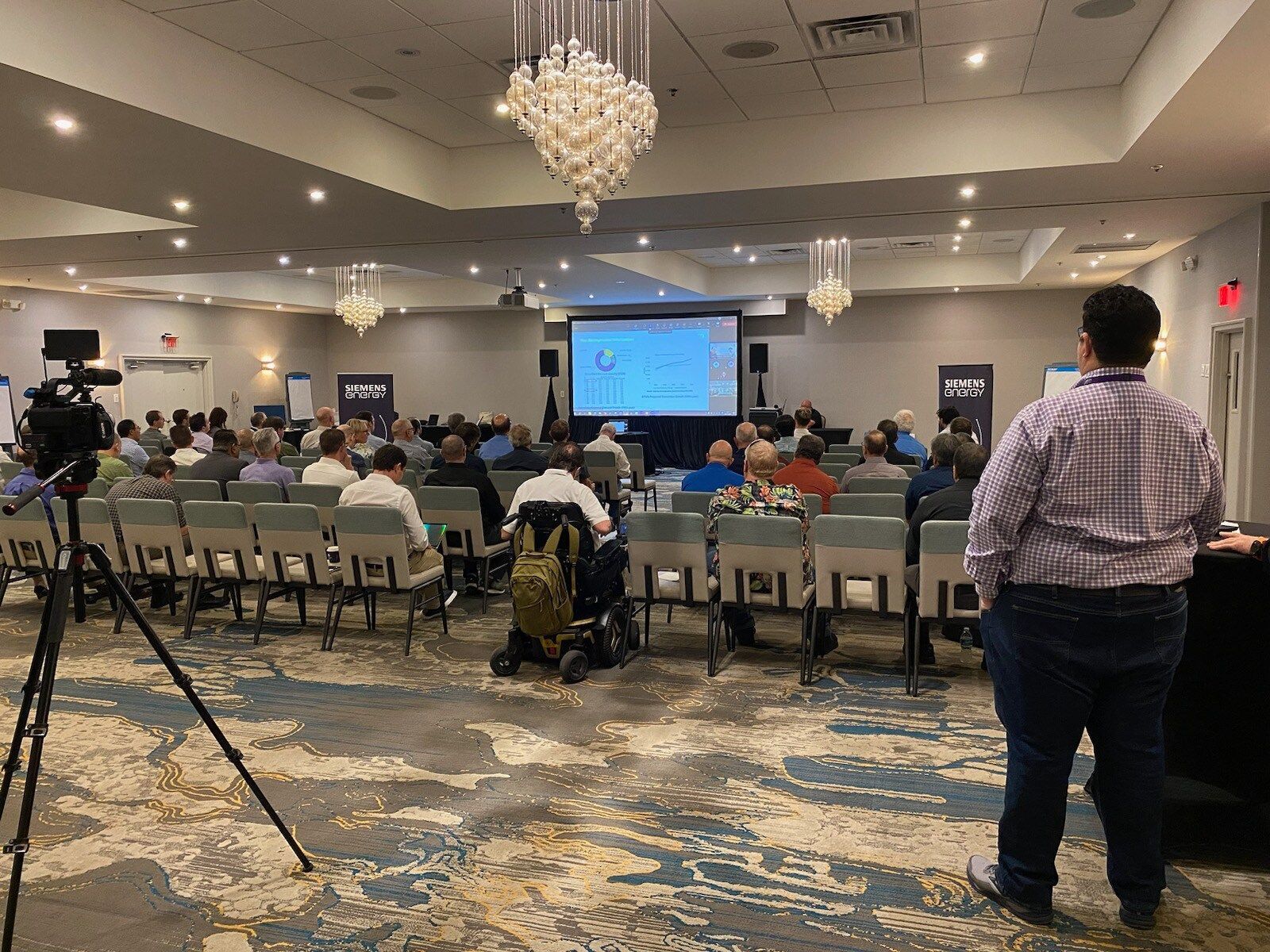 Conference room presentation with seated audience, blue slide screen, and a man standing at the right.