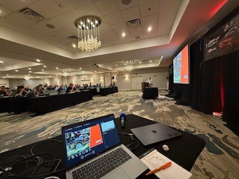 Conference room with laptop at podium, projection screen, and attendees seated at long tables