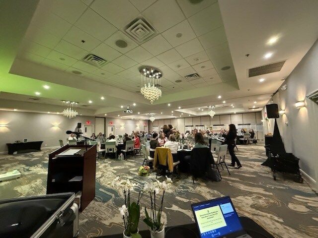 Conference room with people seated at round tables under chandeliers, viewed from a podium.