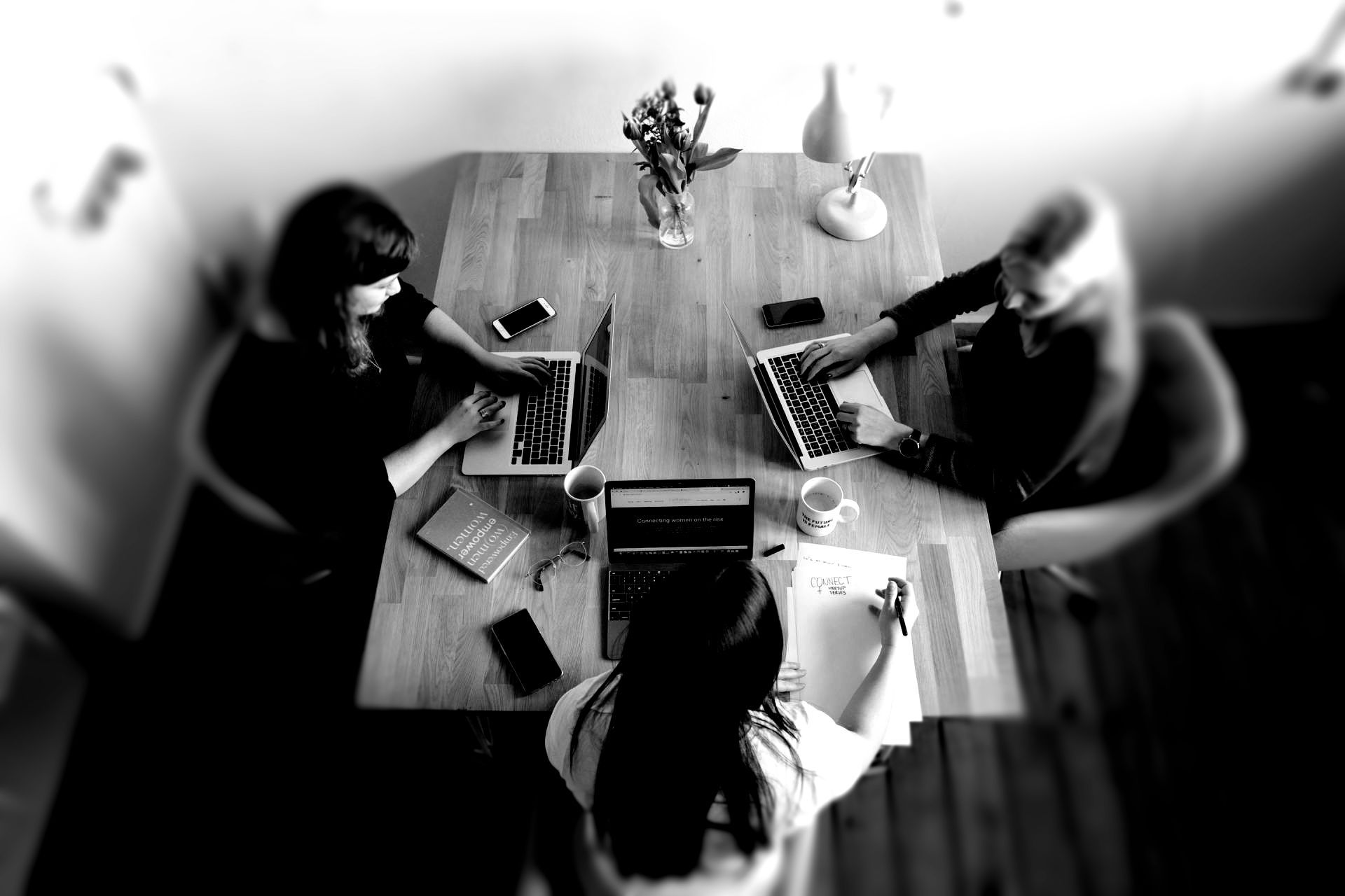 Three people work on laptops around a wooden table in a black-and-white overhead view.