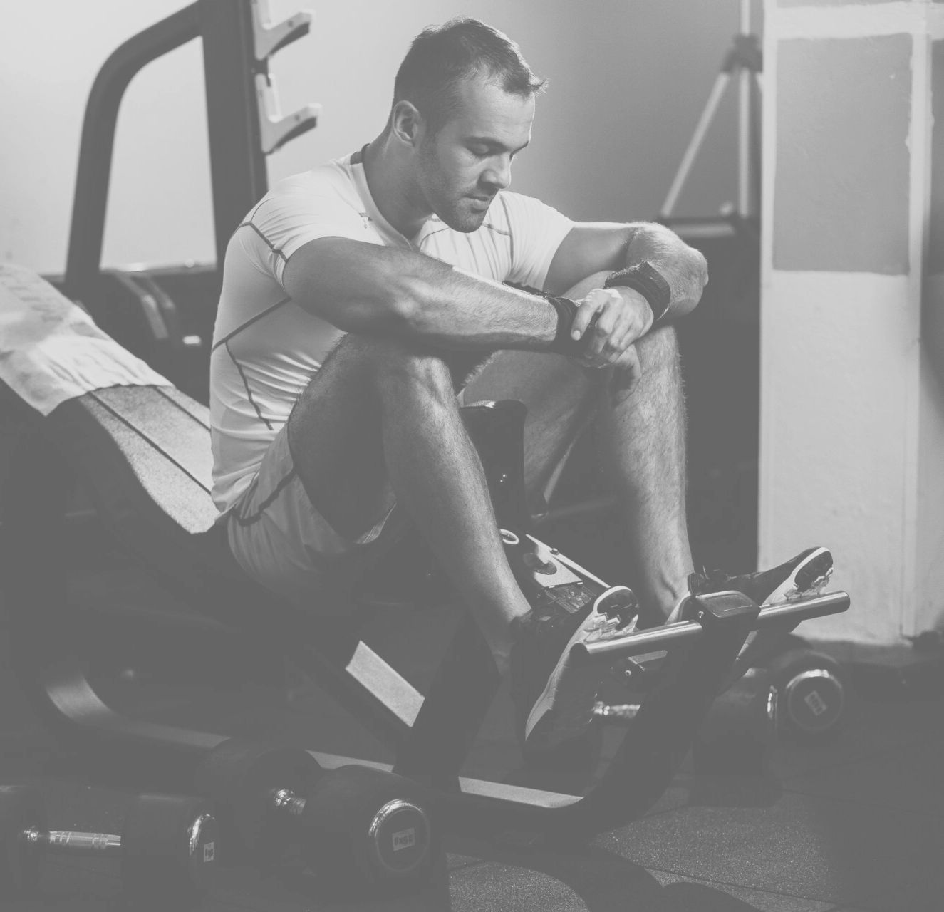 Man sitting on a gym machine, resting his arms on his knees in a monochrome gym scene