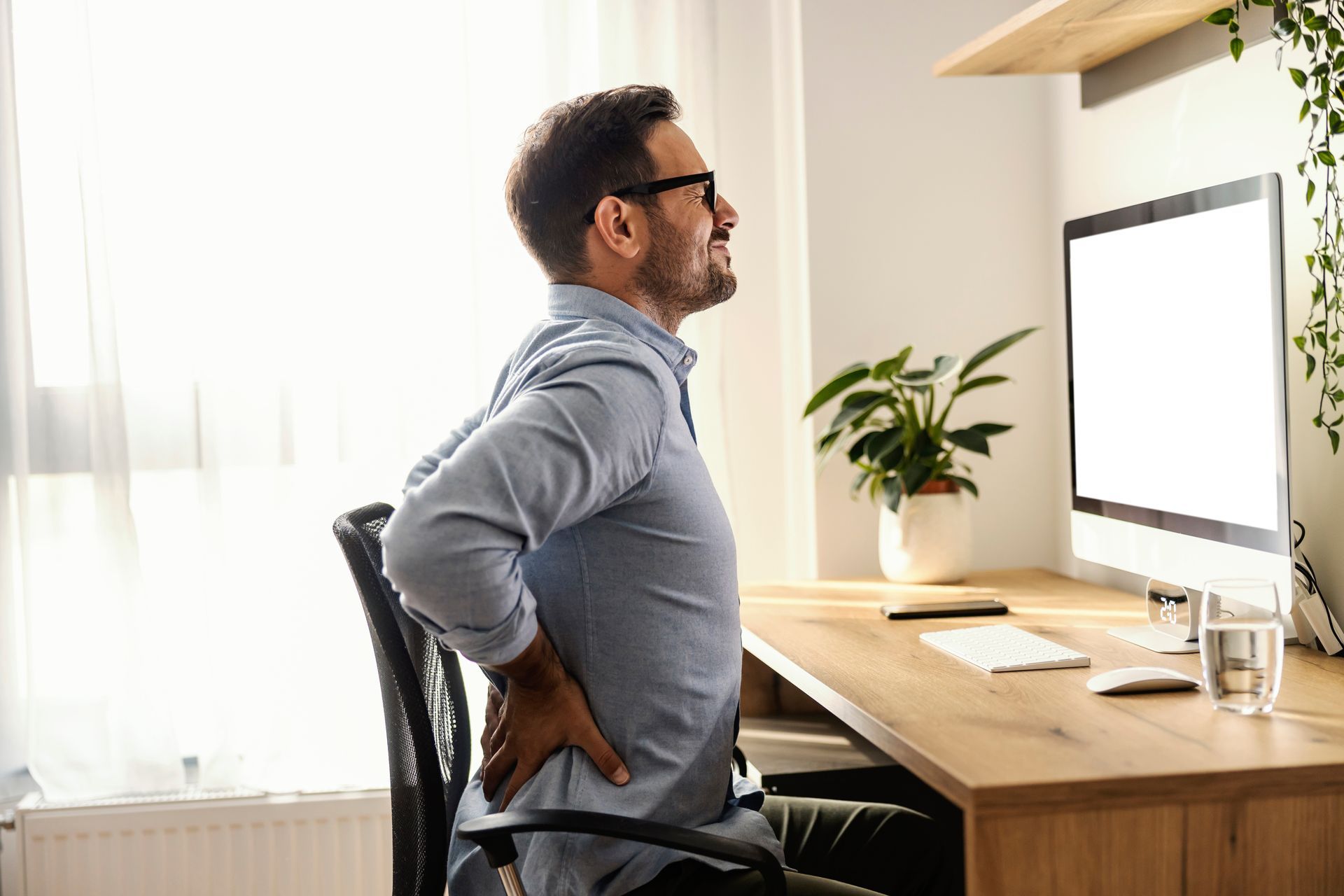 Man at a desk holding his lower back in pain while working on a computer.