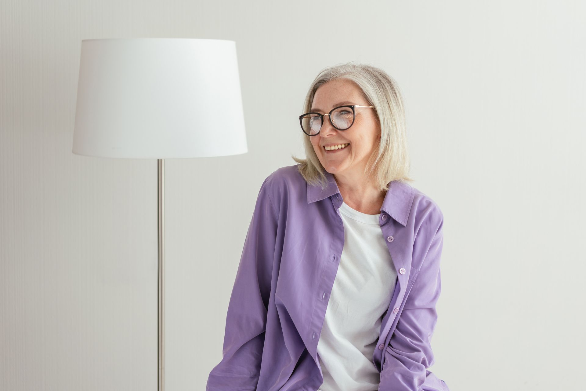 Eine Frau im besten Alter mit Brille und lila Hemd sitzt vor heller Wand neben einer hellen Lampe.