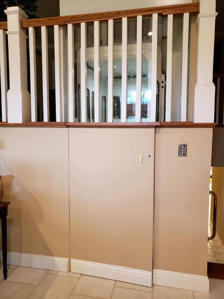 A tan-colored wooden elevator shaft with a closed door sits beneath a white banister and wooden railing in a home.