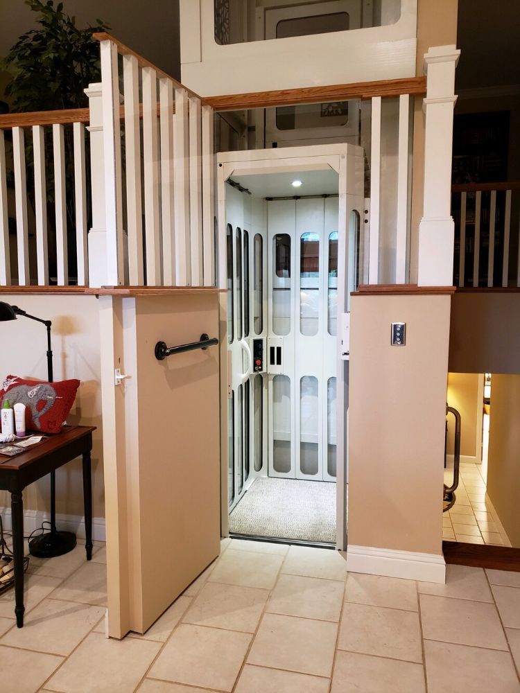 A residential elevator with white, framed glass doors centered between beige walls and wooden stair railings.