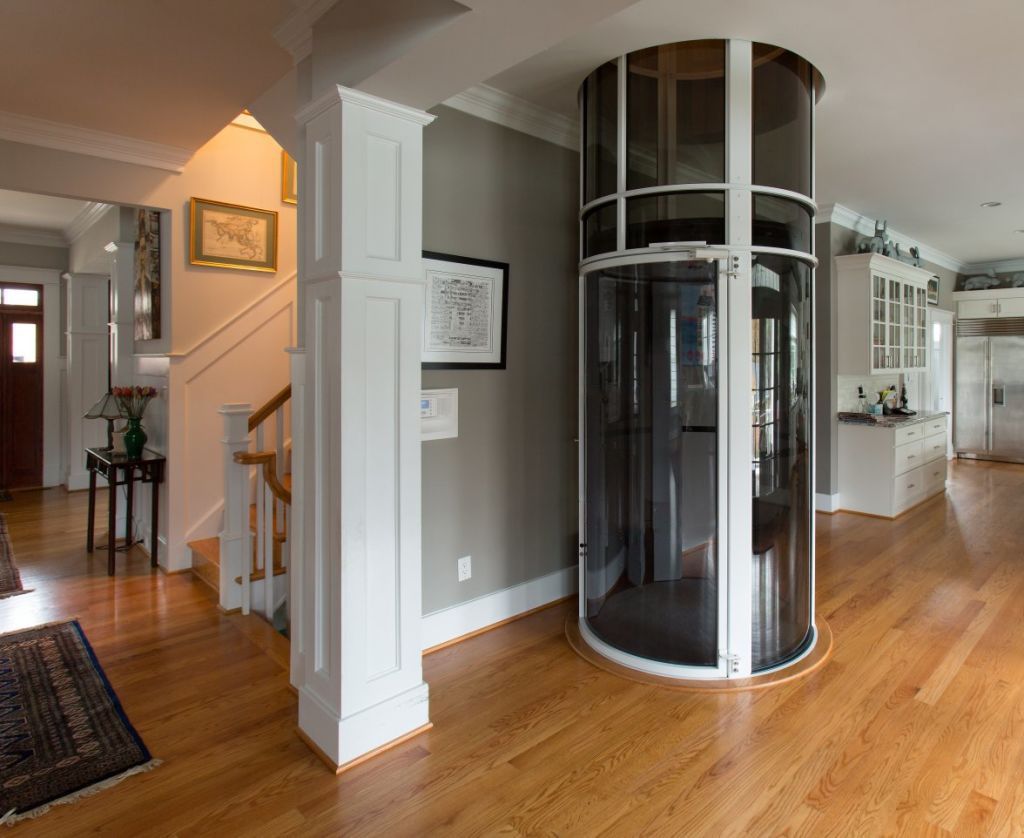 A cylindrical, glass-walled residential elevator stands on a light hardwood floor next to a white column in a home.