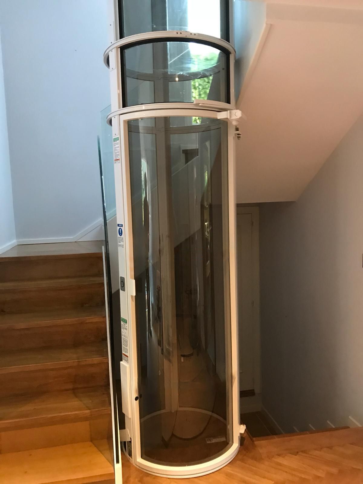 A cylindrical, white-framed glass residential elevator installed between levels of a home with wooden stairs.