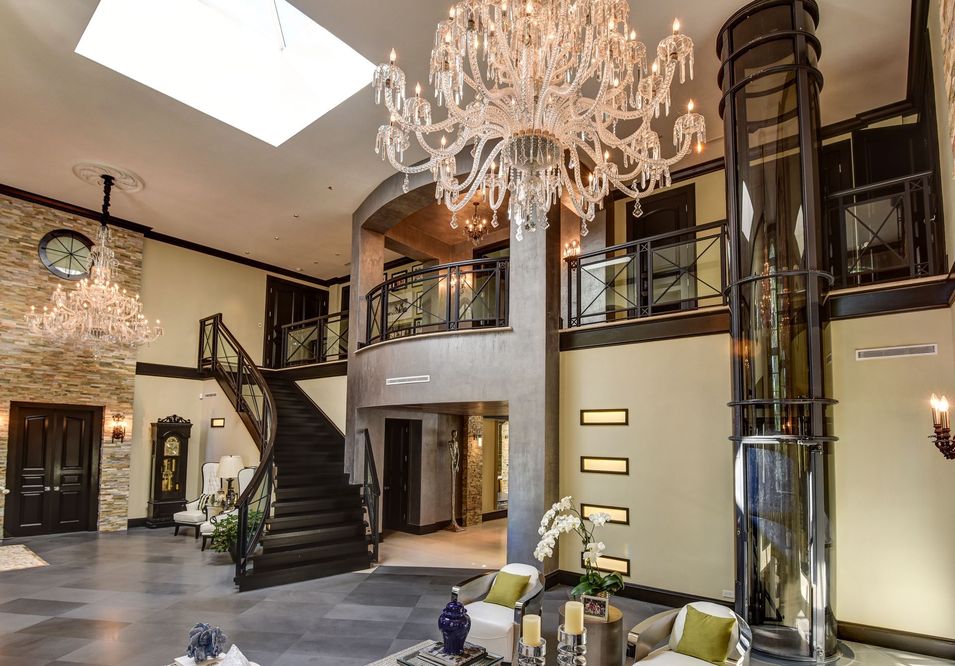 Grand foyer with a vaulted ceiling, crystal chandelier, stone wall, curved staircase, and a cylindrical glass elevator.