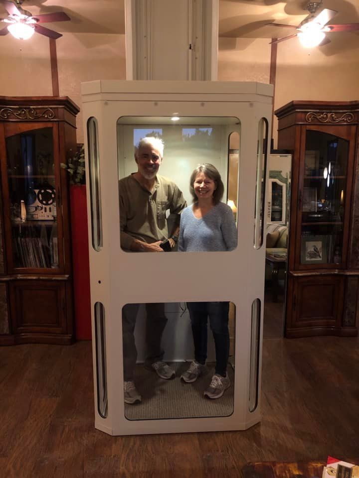 A man and a woman stand inside a white, residential elevator with glass panels, positioned between two wooden cabinets.