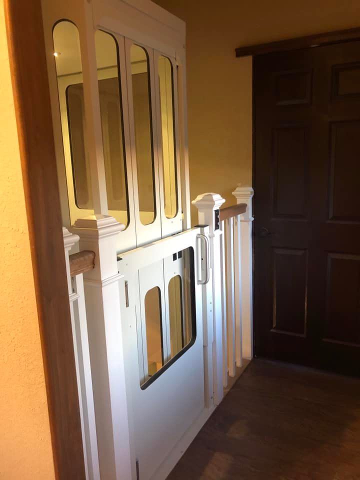 A white residential elevator door with glass panels installed in a home hallway next to a wooden banister and door.