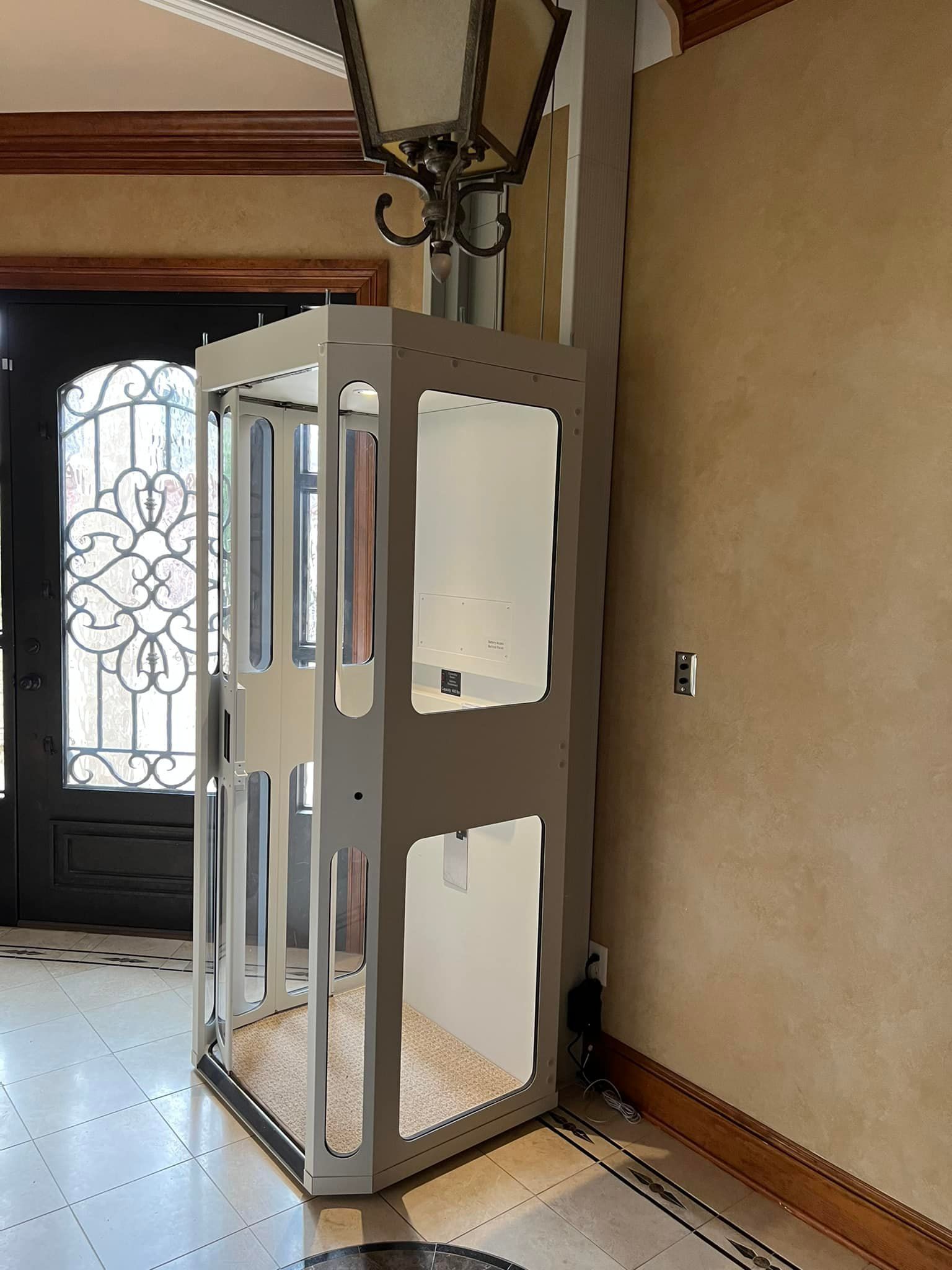 A white, framed residential elevator with glass panels stands in a tile-floored home entry near a decorative front door.