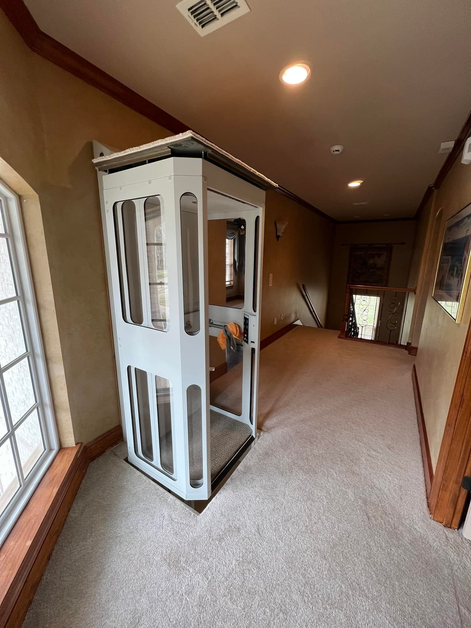 A white, caged residential elevator standing in a carpeted hallway next to a large window.