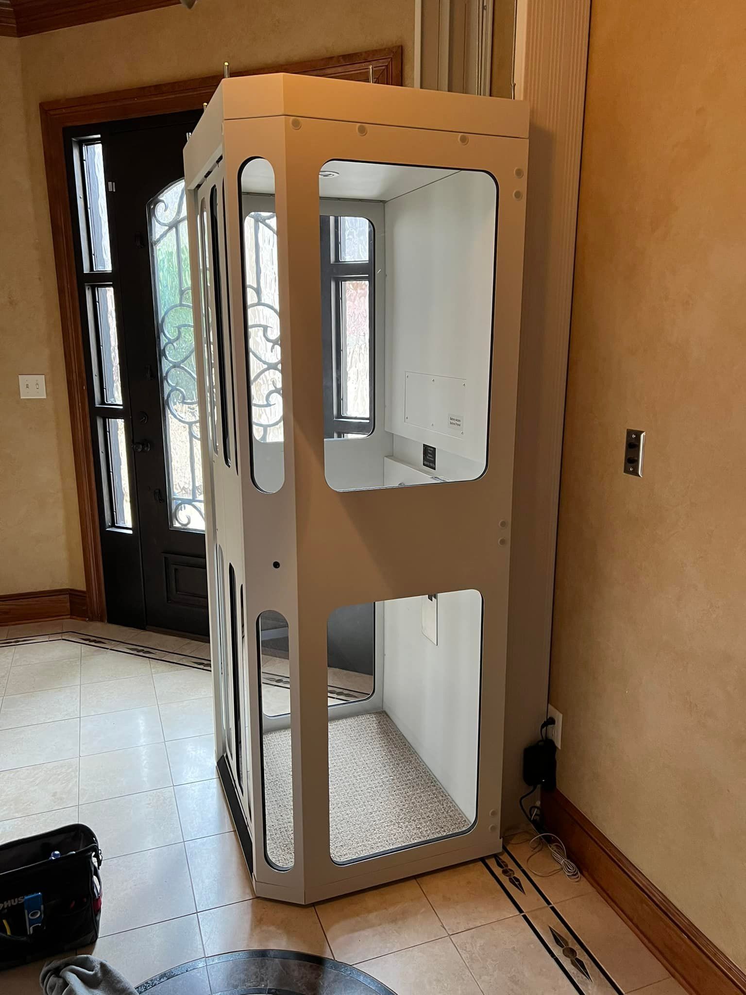 A white, rectangular, modular residential elevator hoistway frame standing in a tiled home entryway.
