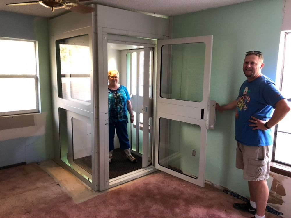 A person stands inside a residential elevator while another person stands next to the open door in a room with carpet.