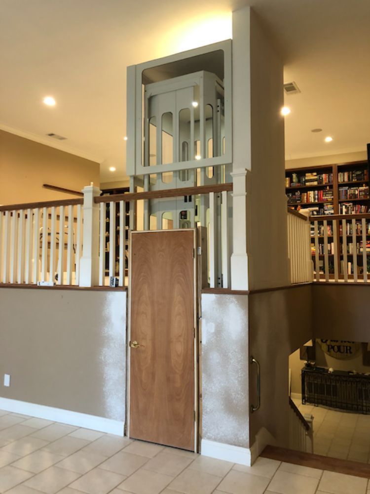A home elevator with glass panels stands centered behind a wooden door, surrounded by an indoor balcony and staircase.