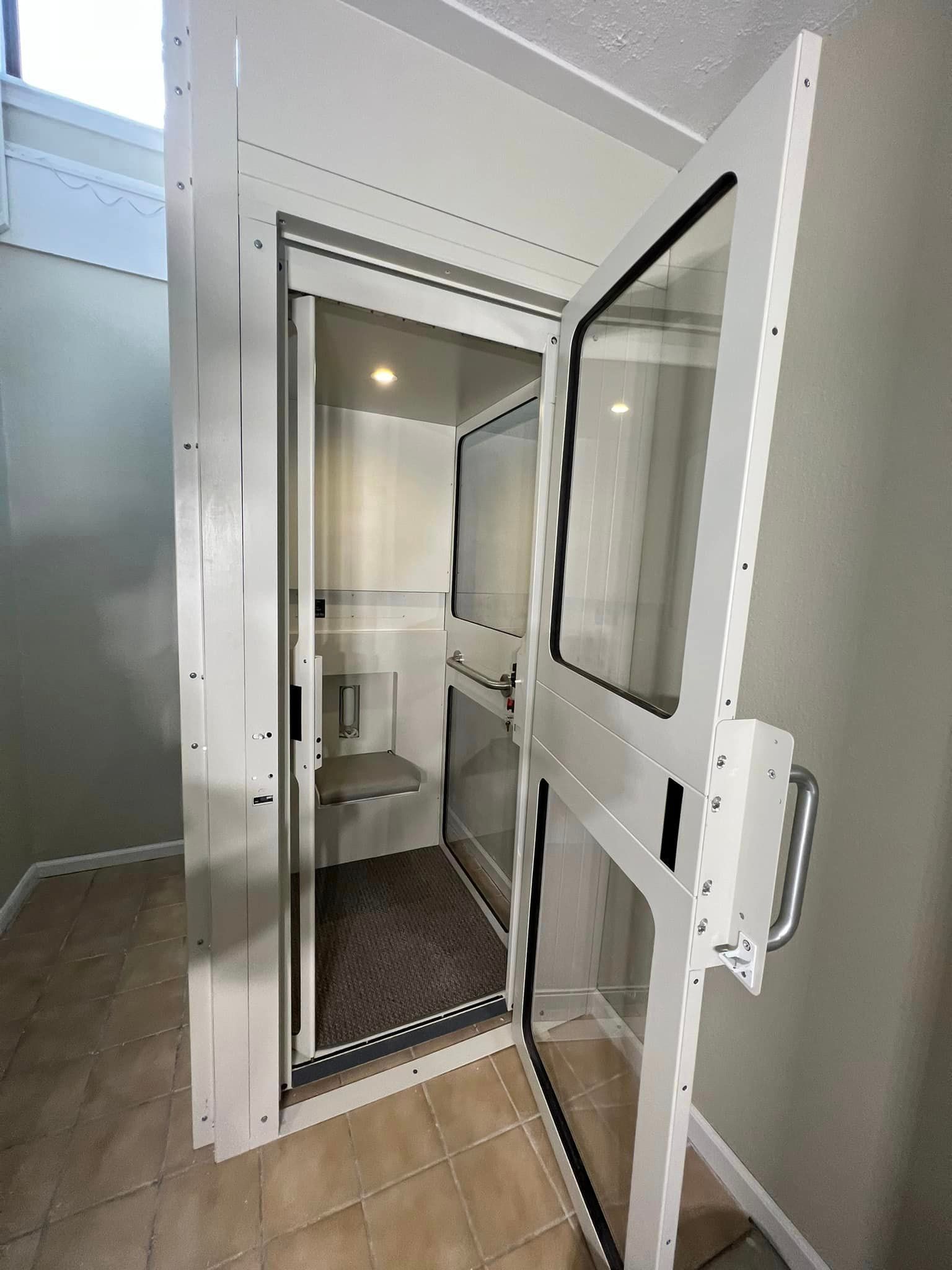 A white residential home elevator with a glass-paneled door, open to reveal a carpeted interior and a small wall seat.
