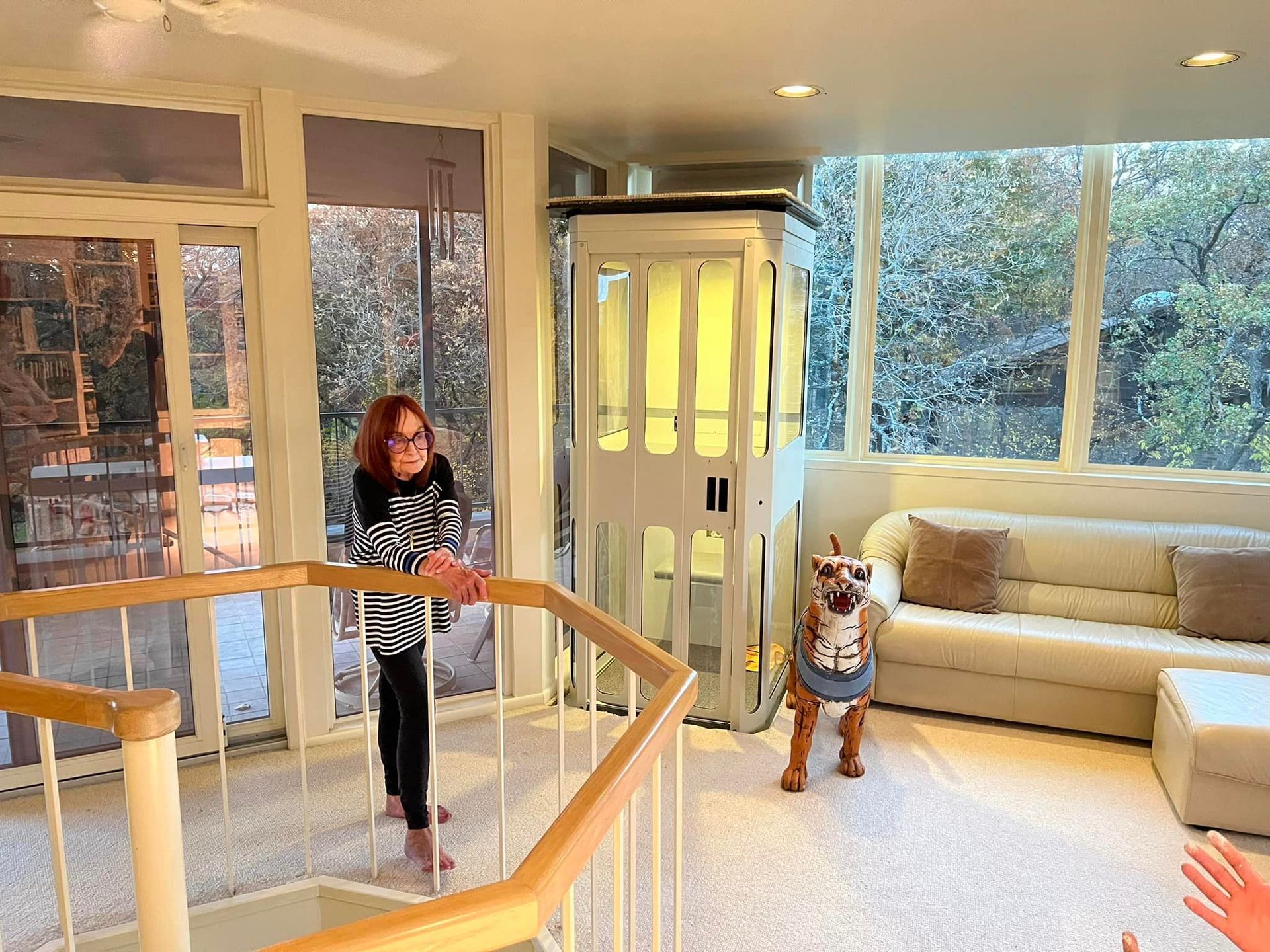 A person stands near a wooden staircase railing in a sunroom next to a white home elevator and a dog on a light carpet.