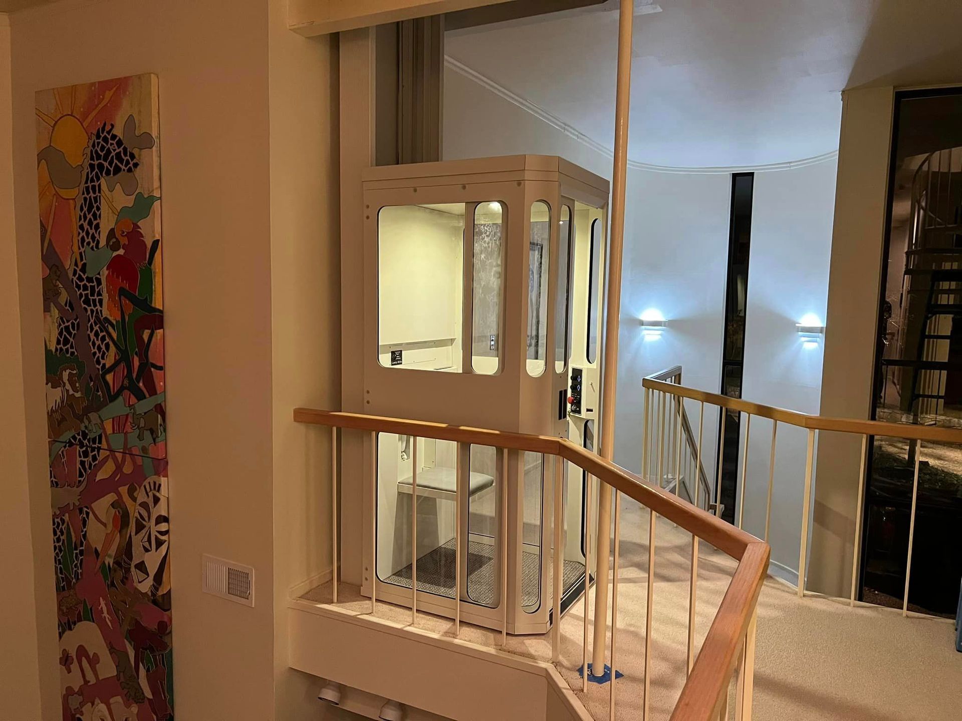 A white residential elevator sits on a carpeted floor next to a wooden railing and a tall, colorful giraffe painting.