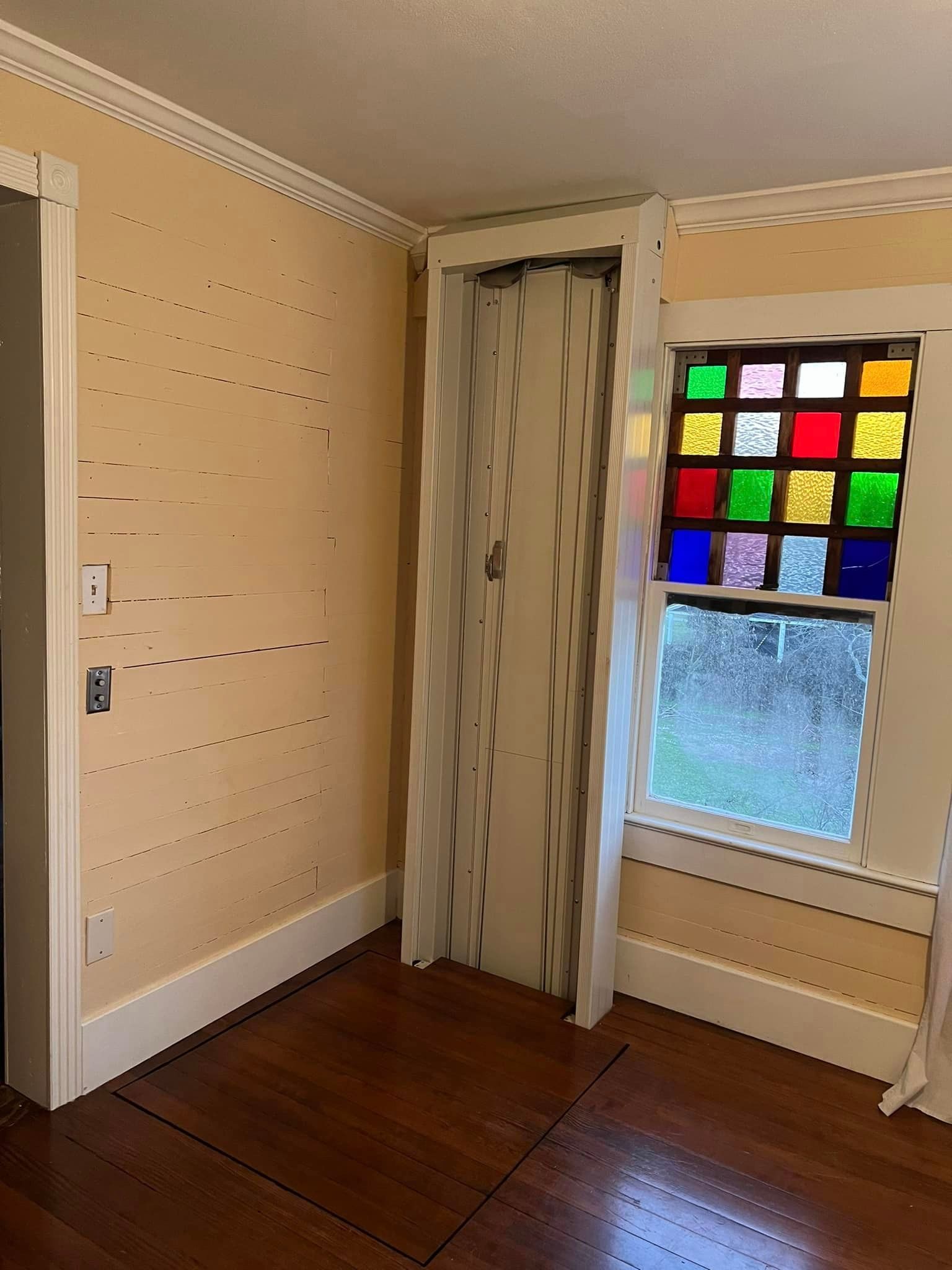 An indoor room with beige, horizontally lined walls, wooden floors, a folding door, and a colorful grid-patterned window.