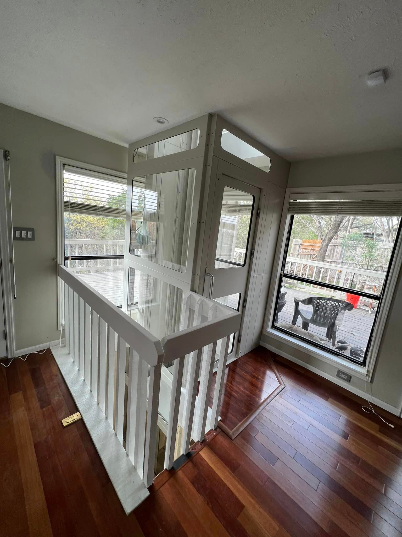 A residential vertical wheelchair lift installed indoors next to a wooden staircase railing in a room with hardwood floors.