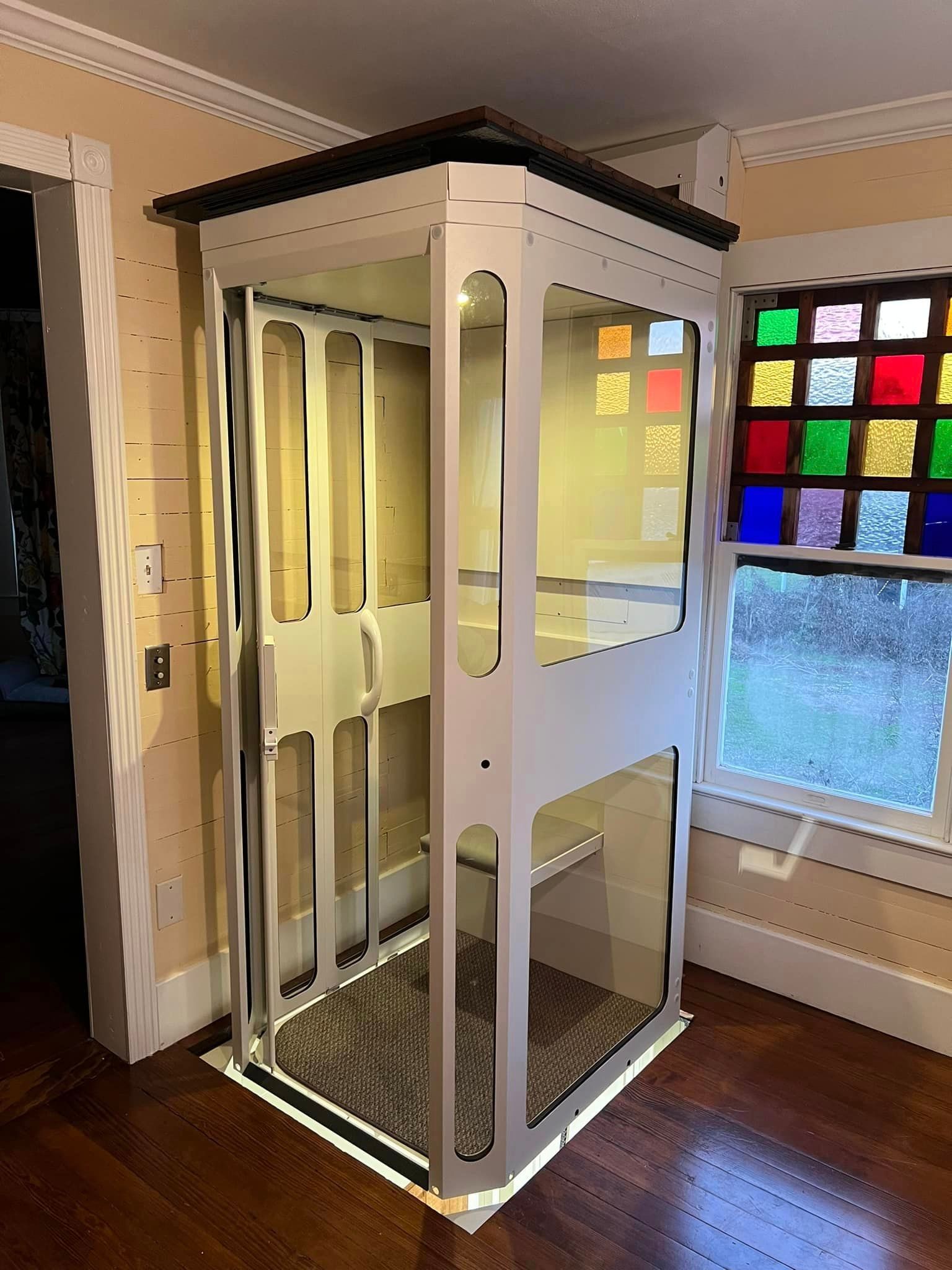 A white home elevator with glass walls installed in a room with hardwood floors and a colorful stained glass window.