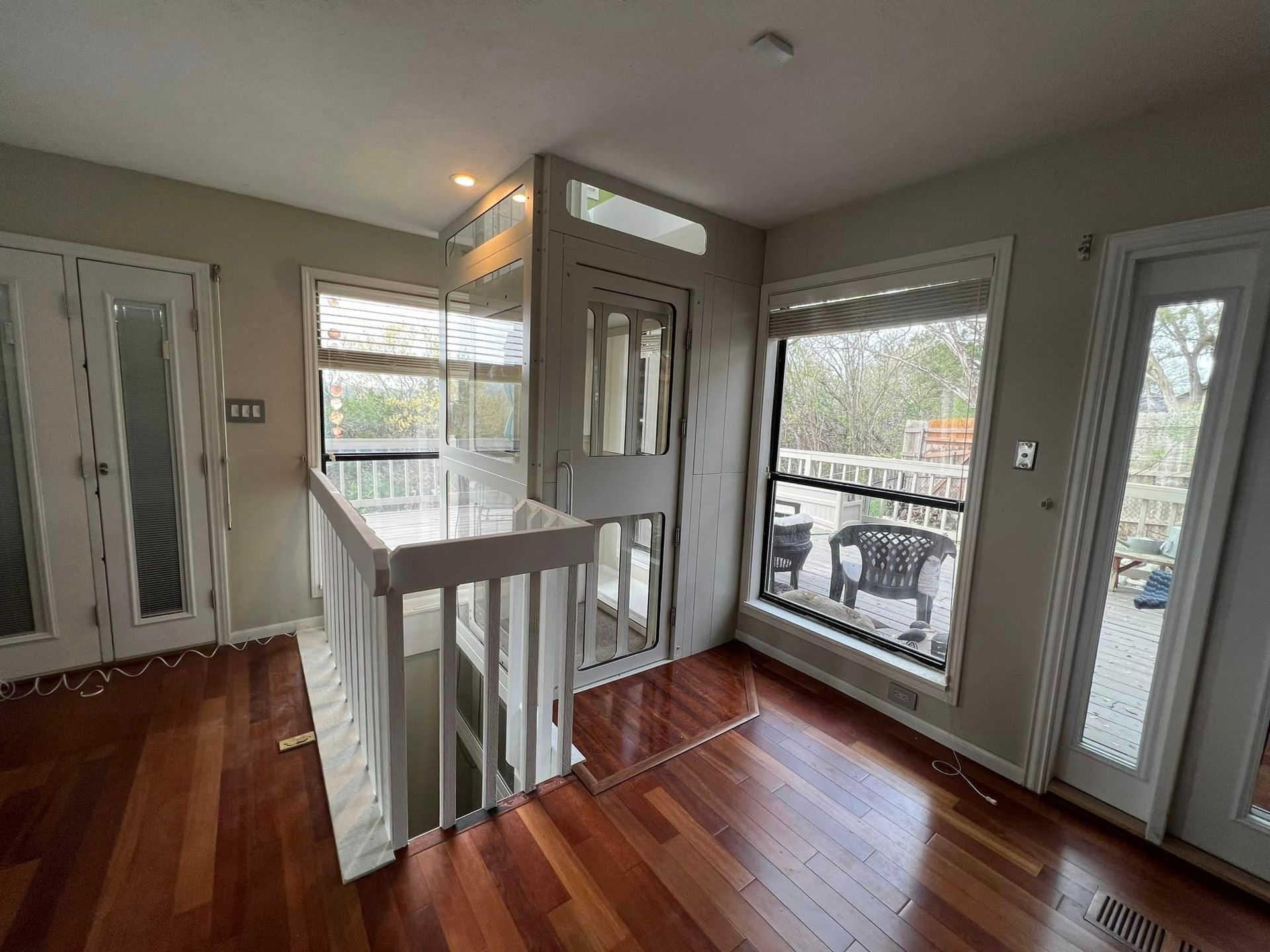 A residential space featuring a floor-to-ceiling glass elevator, white staircase railings, hardwood floors, and a balcony.