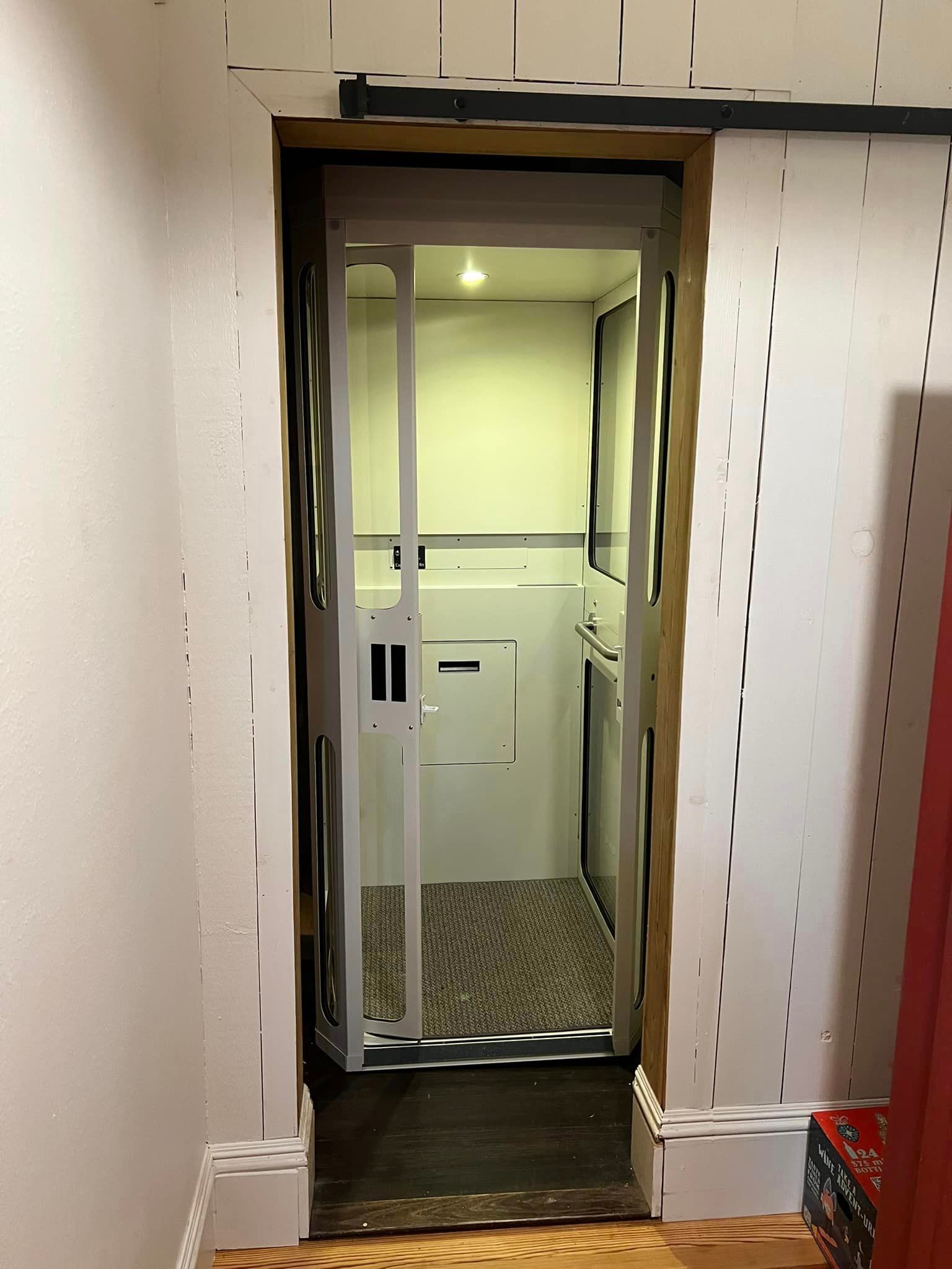 An interior home elevator with an open folding gate, set within a residential hallway with light walls and dark flooring.