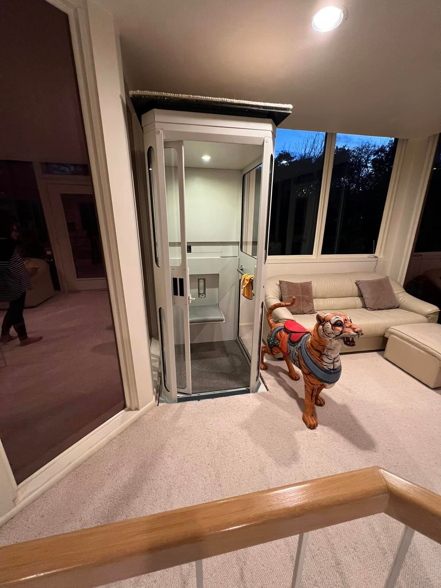 A residential elevator with a built-in seat stands in a room with a sofa and a large tiger statue on the carpet.