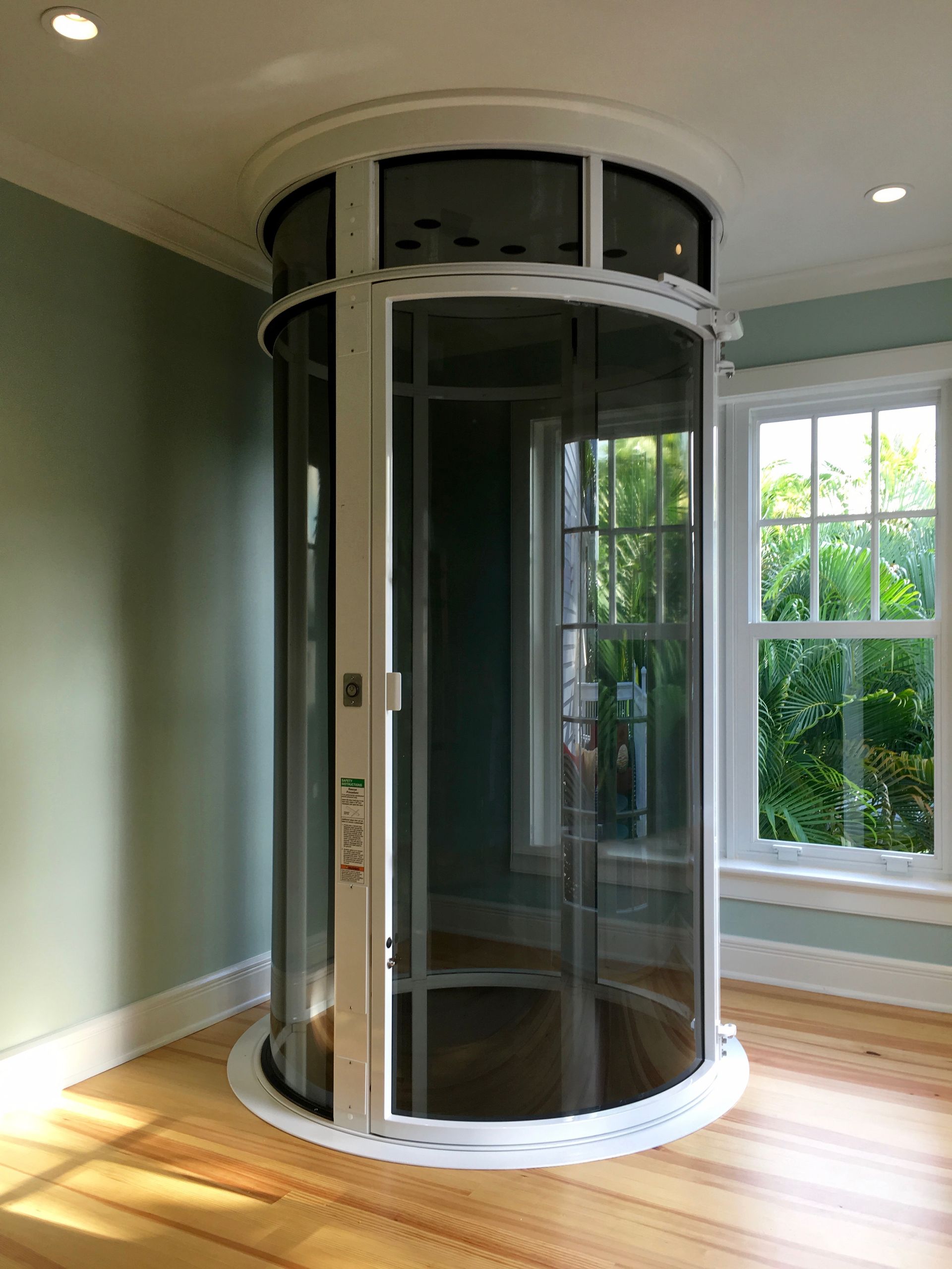 A residential vacuum elevator with a white frame and curved glass panels stands in a room with hardwood floors.