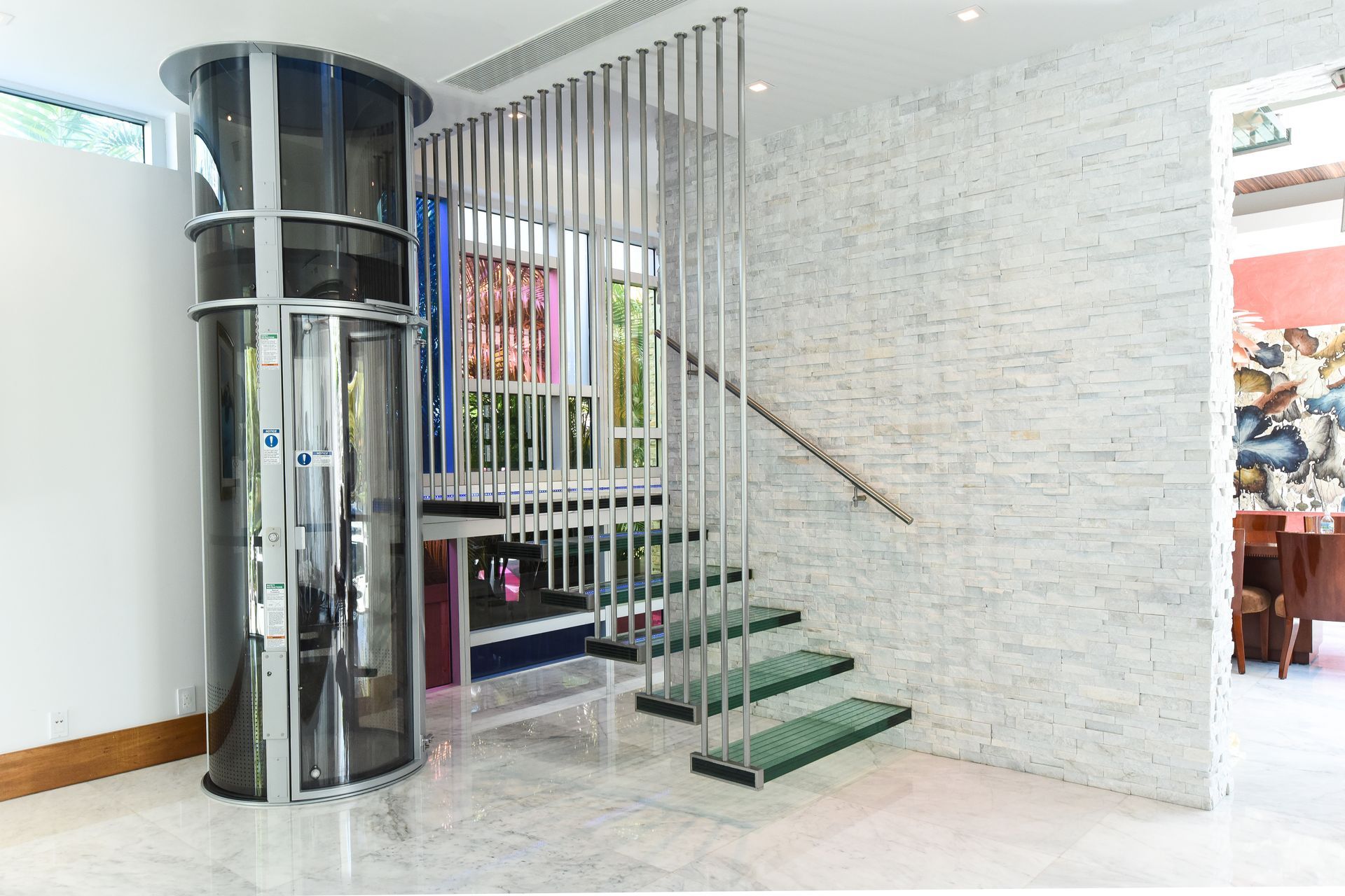 A circular glass elevator stands next to a modern staircase with floating green glass steps and a vertical metal railing.