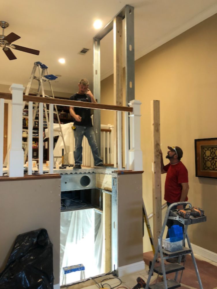 Two workers install a metal frame wall partition on a landing overlooking a staircase.