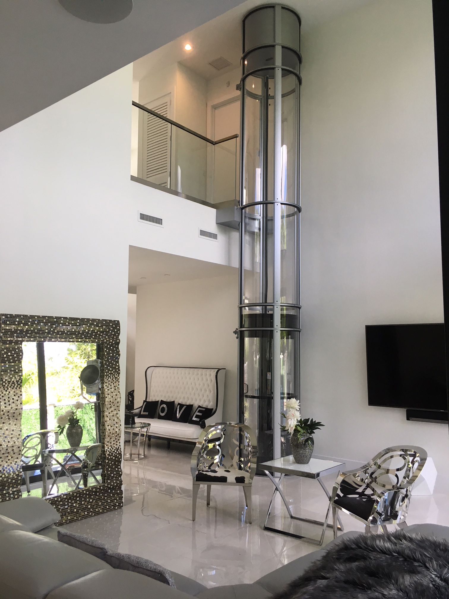 A cylindrical, glass-walled residential elevator stands in a modern, white living room with high ceilings and a balcony.