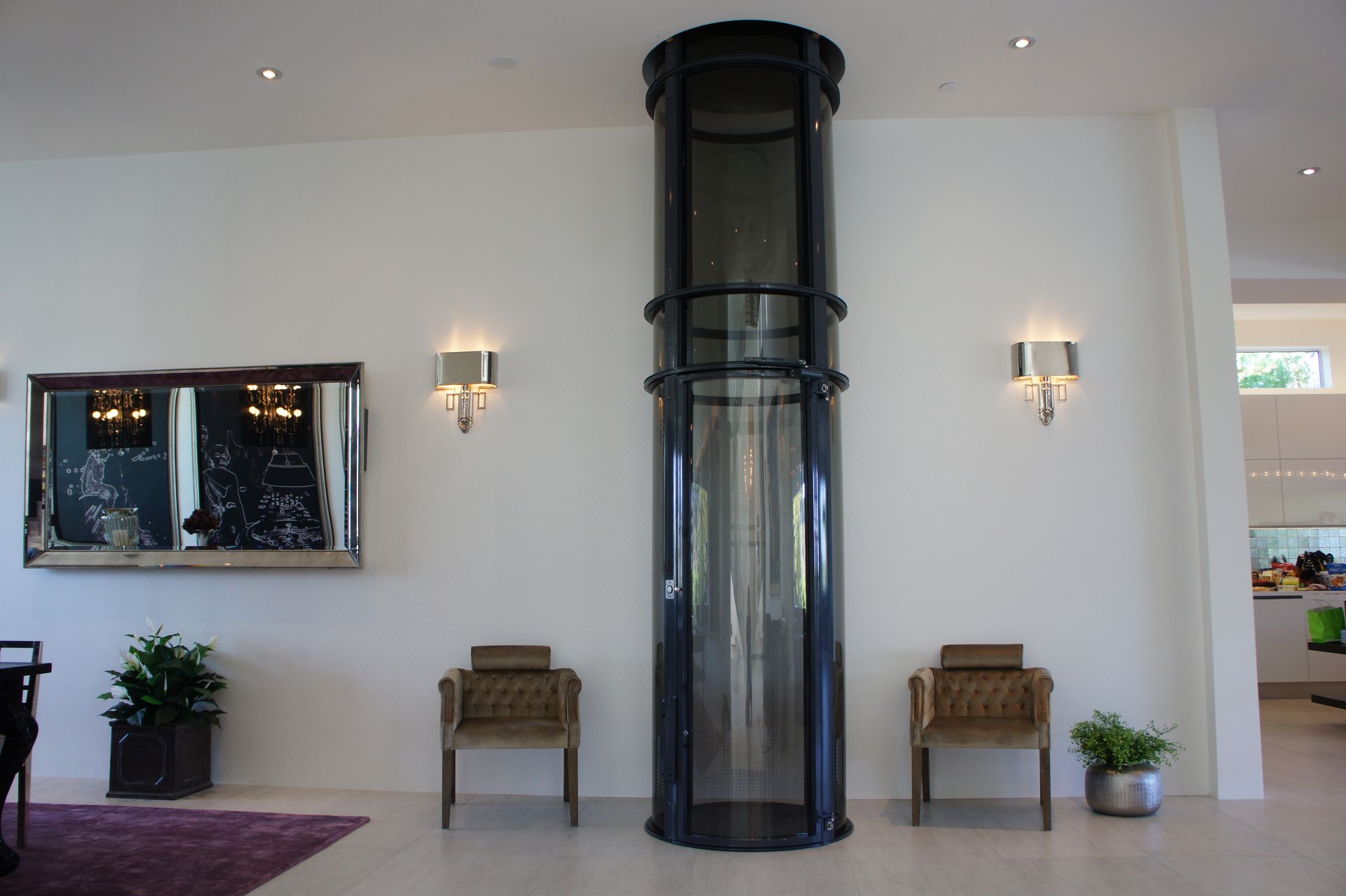 A modern, cylindrical glass elevator stands between two armchairs against a white wall in a bright, minimalist room.