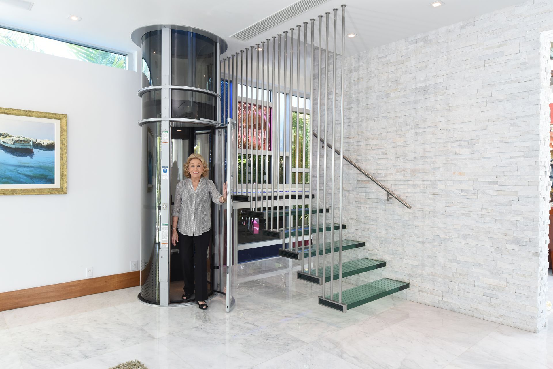 A person stands inside a sleek, cylindrical glass home elevator next to a modern staircase with floating glass steps.