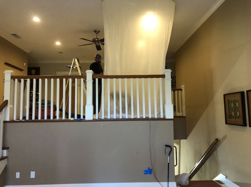 A worker stands behind a white plastic barrier on an indoor balcony overlooking a staircase in a house.