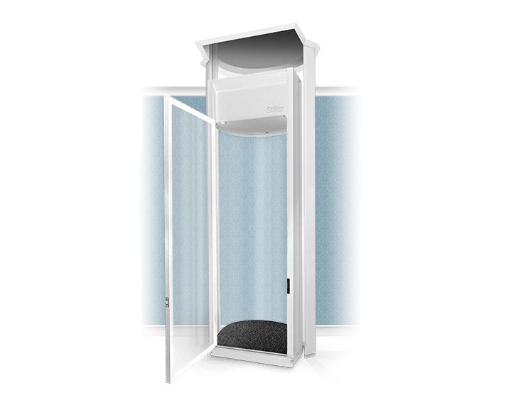 A white home elevator installed in an interior hallway, adjacent to a wooden staircase, a lamp, and a potted plant.