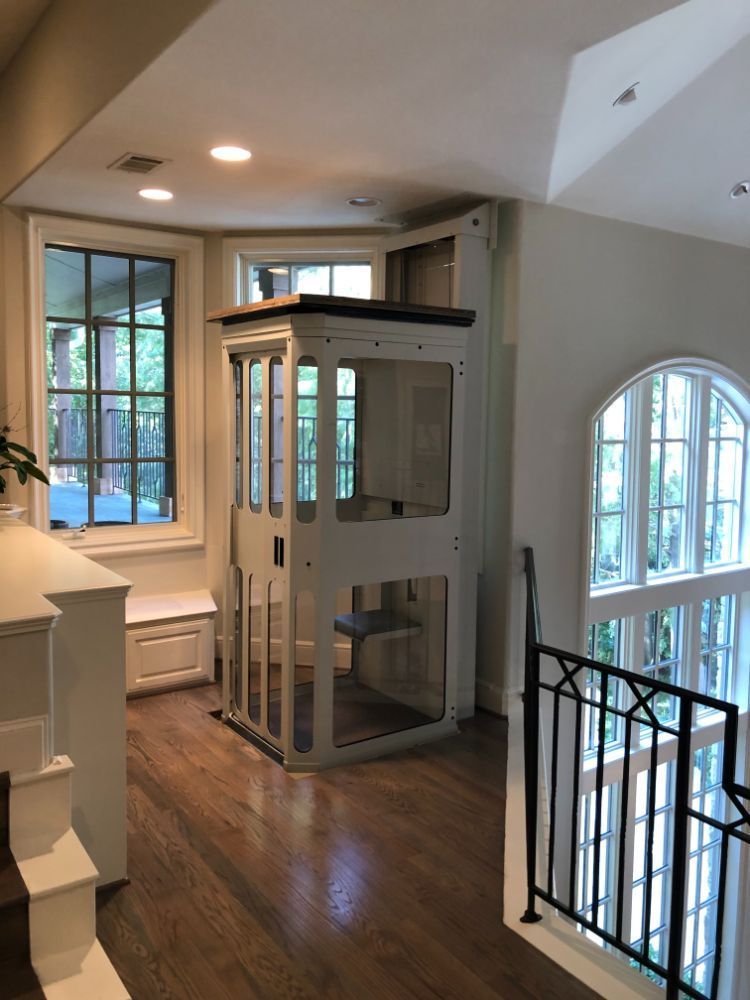A residential elevator enclosed in glass and light-colored wood, positioned on a wooden floor in a home interior.