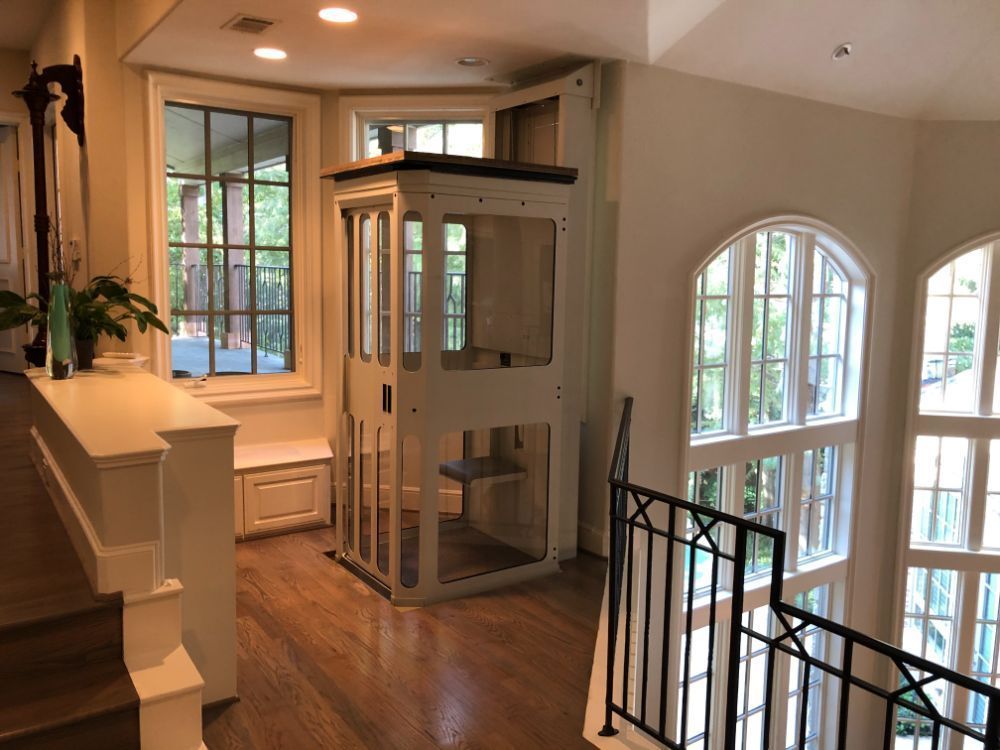 A white residential elevator stands in a light-filled home hallway next to a staircase and large windows.