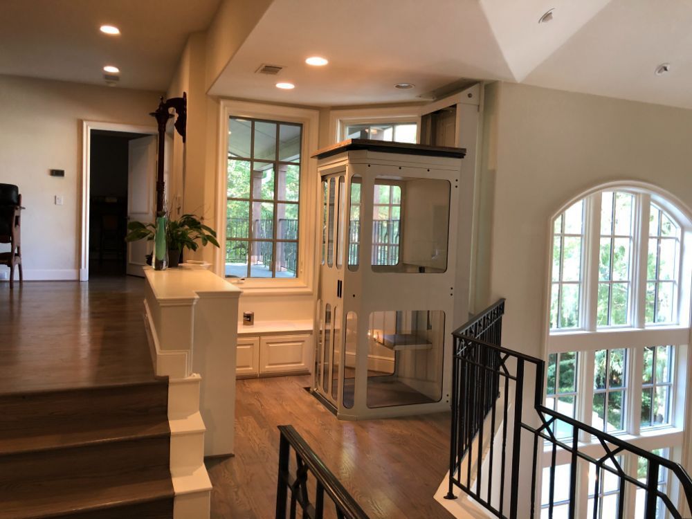 A white, residential home elevator stands next to a large windowed alcove at the top of a wooden staircase.