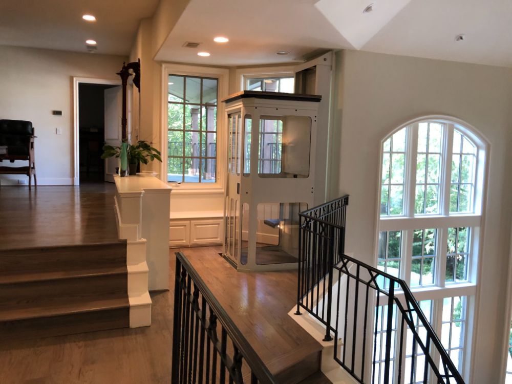 A home elevator installed in an open landing area next to a wooden staircase, large windows, and black metal railings.