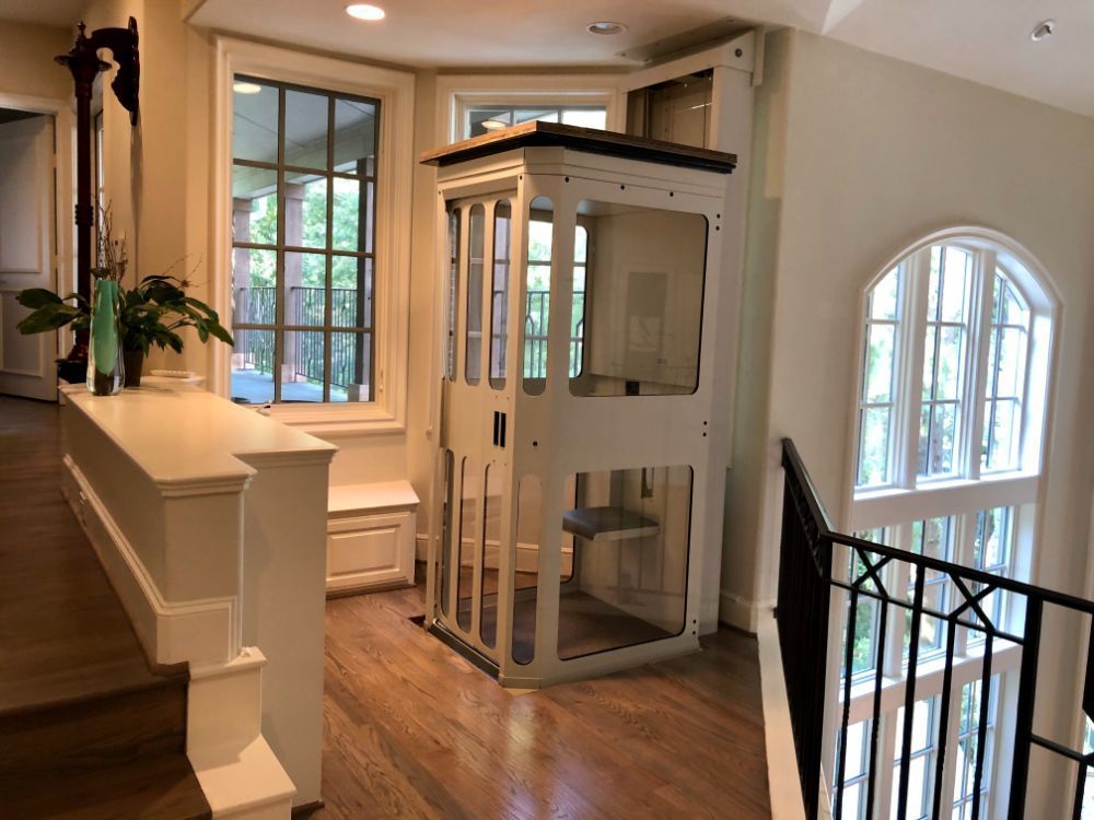 A white residential elevator stands in a bright, wood-floored home hallway near a staircase and windows.