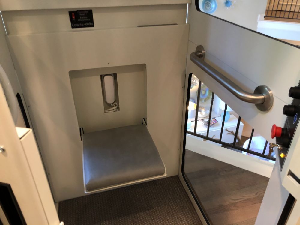 A vertical wheelchair lift interior showing a fold-down seat, grey flooring, a metal handrail, and an emergency stop button.