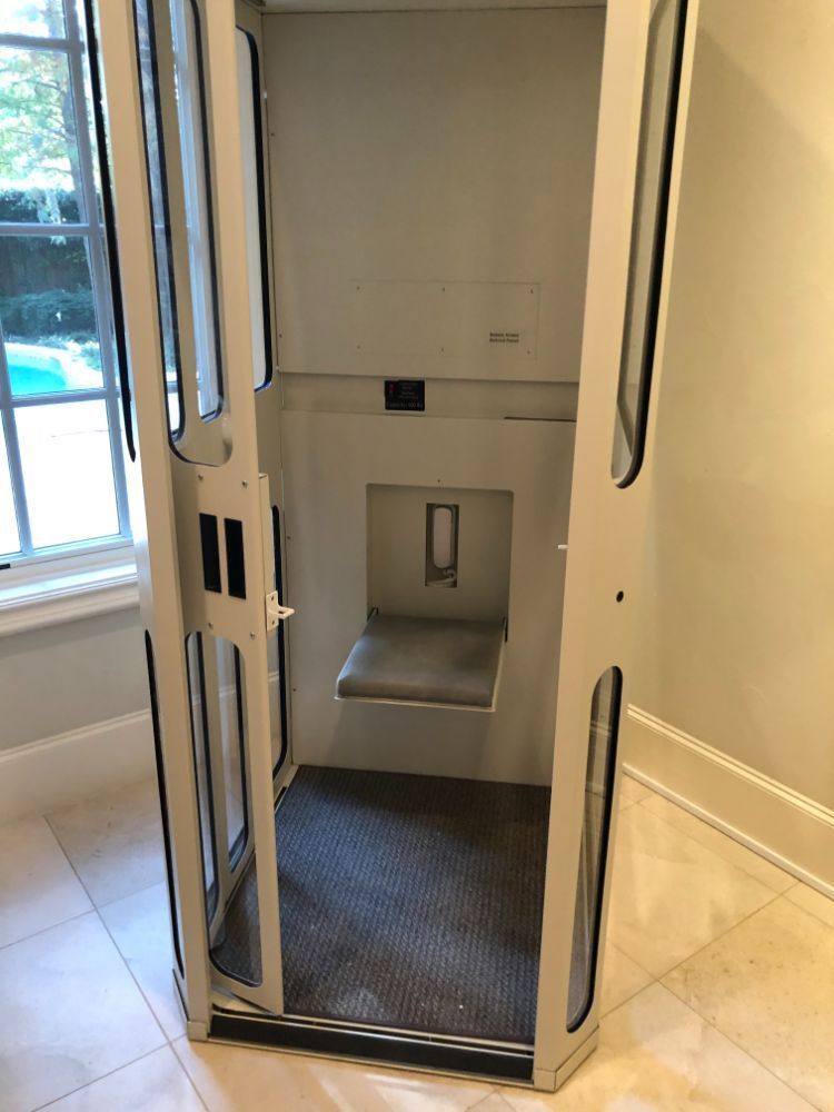 A residential elevator with a small, fold-down seat inside a white frame with glass panels, situated in a tiled room.