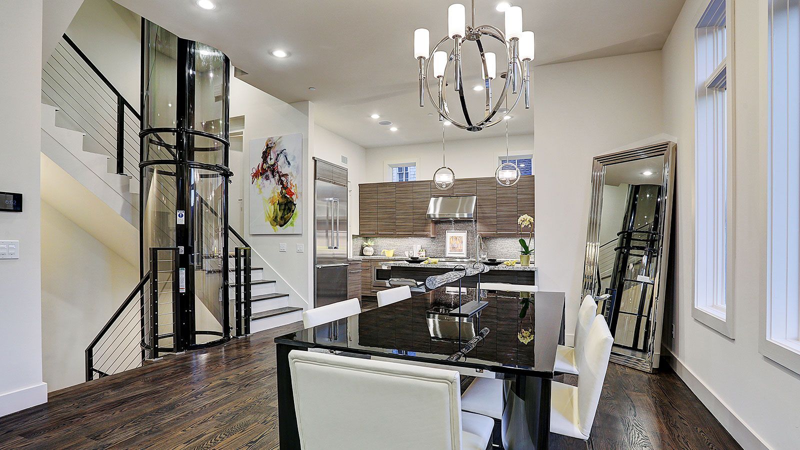A modern dining area with a glass elevator, dark wood floors, a black table, and a large ornate floor mirror.