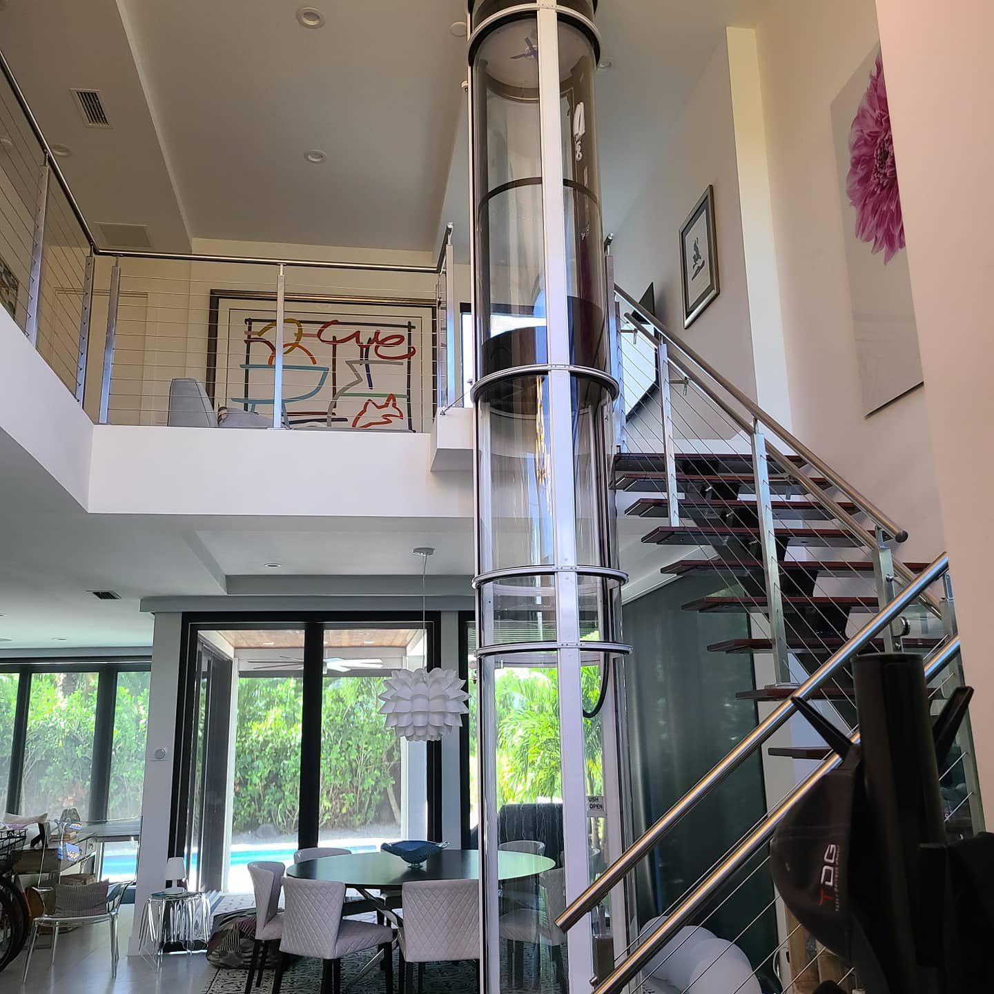 Modern home interior featuring a cylindrical glass elevator centered next to a staircase and dining area.