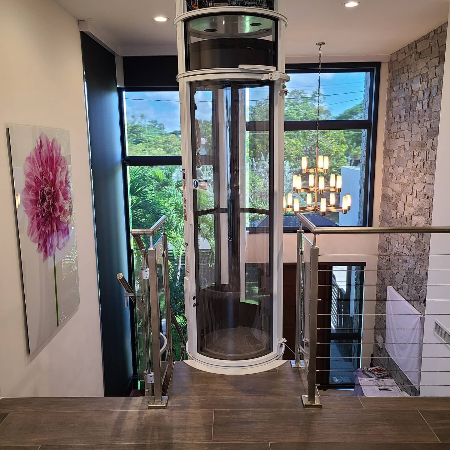 A modern, cylindrical glass home elevator stands in a light-filled entryway between a staircase and a stone wall.
