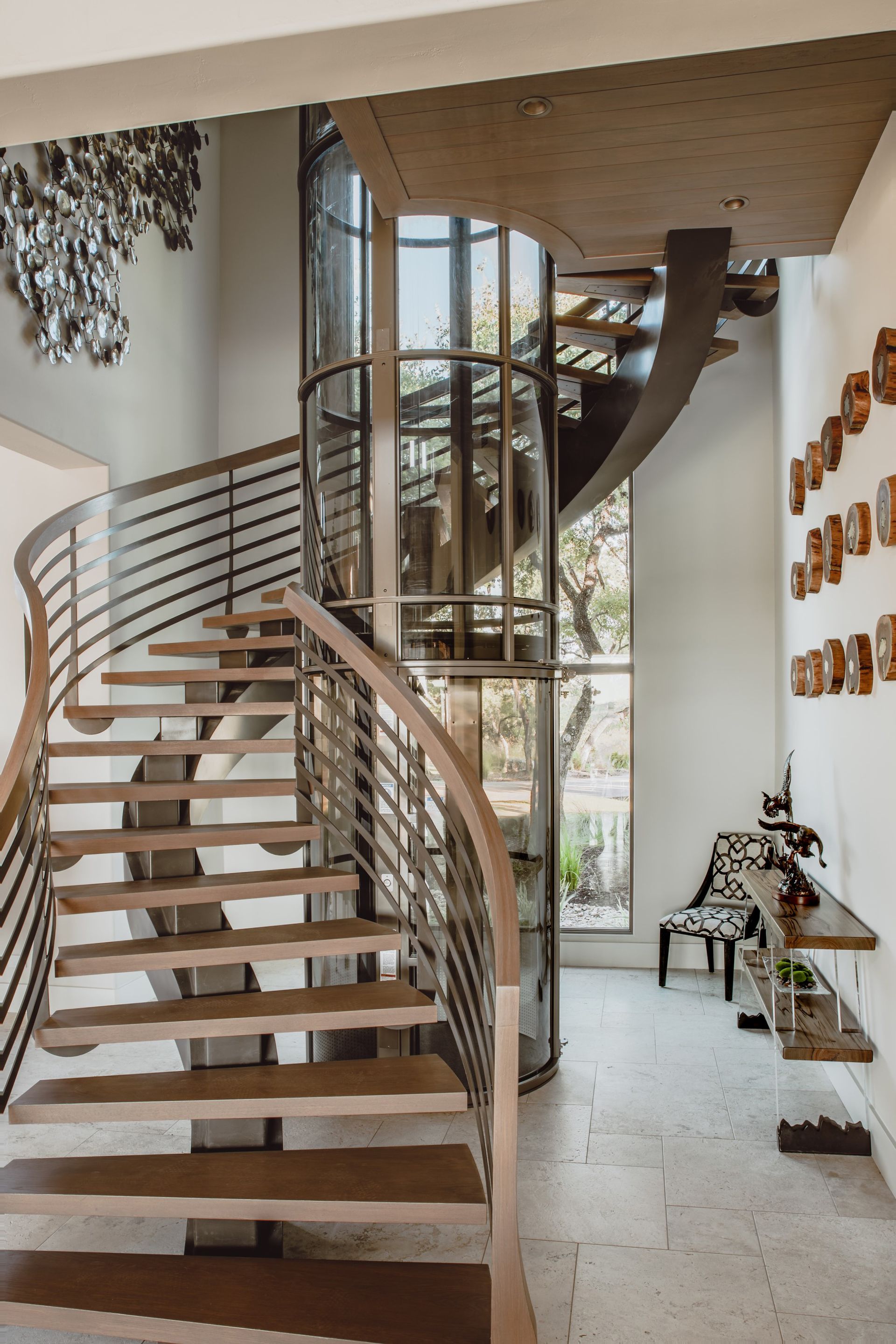 A modern spiral staircase surrounds a cylindrical glass elevator in a bright, contemporary entryway with stone floors.
